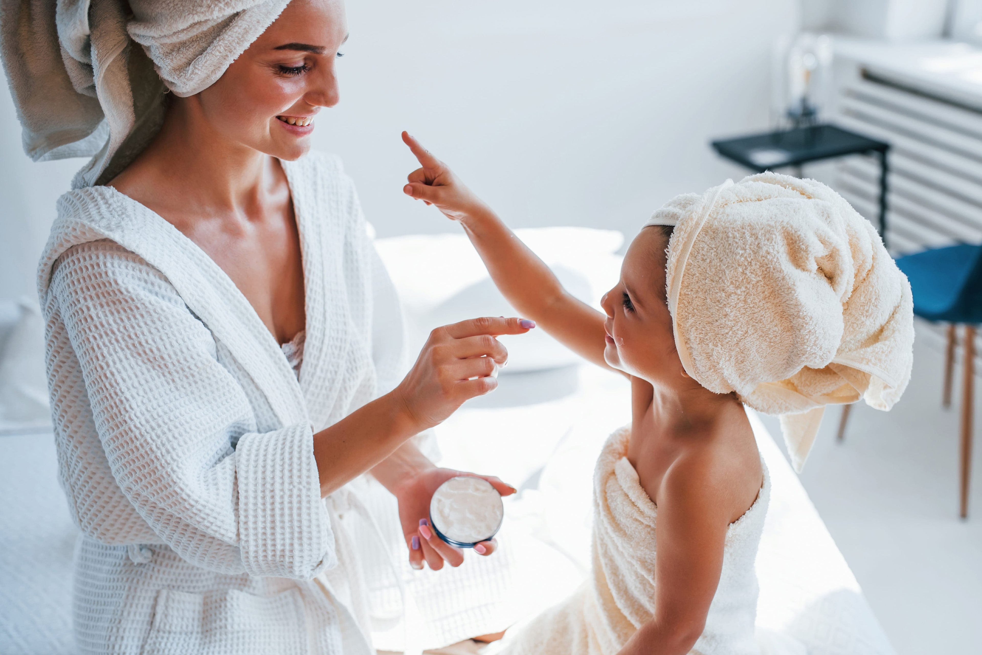 Woman in a bathrobe applying cream to a child's hand with a towel on their head in a bathroom.