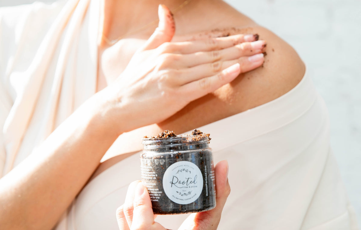 A person wearing a white robe applies a dark brown scrub from a small jar labeled 'Rested' to their shoulder. The product is shown in use in a bright, clean setting.